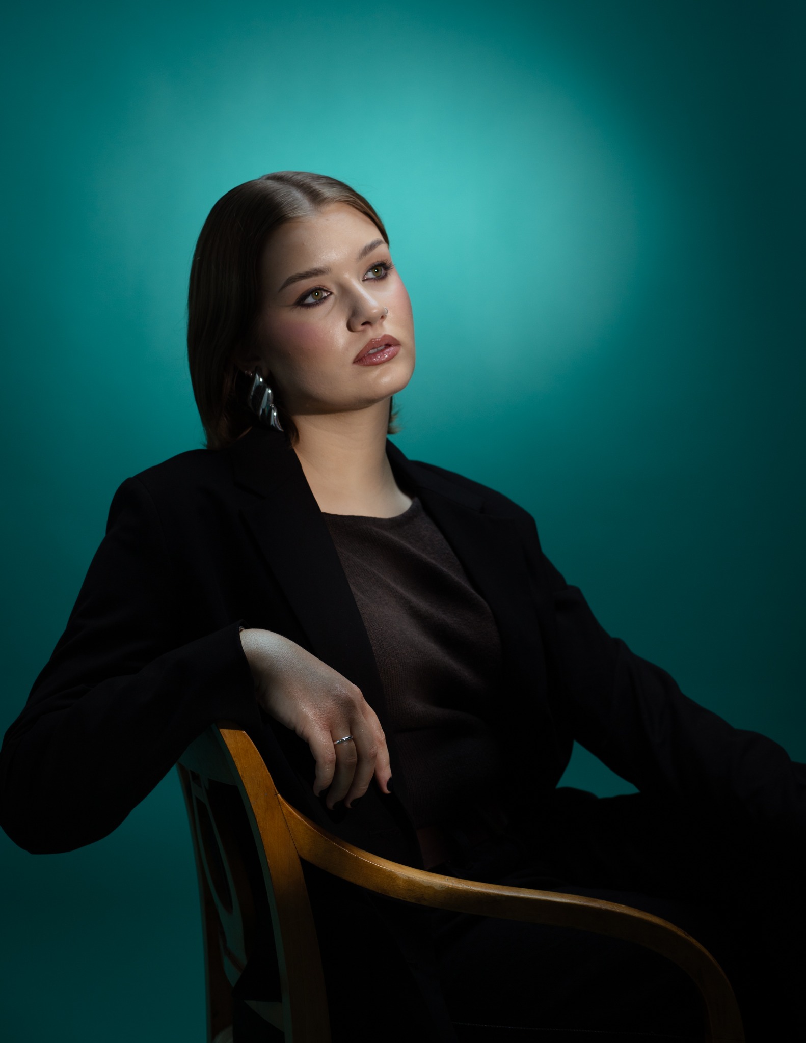 Portret kobiety w czarnym żakiecie, siedzącej na drewnianym krześle na tle turkusowego tła, z delikatnym oświetleniem podkreślającym makijaż i srebrne kolczyki.