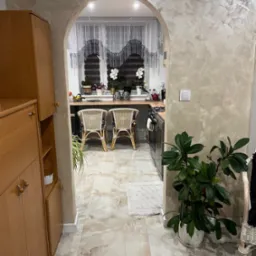 Arched doorway leading to a bright kitchen with textured walls, marble-patterned floor tiles, and a view of wicker chairs at a countertop, accented by indoor plants and decorative curtains.