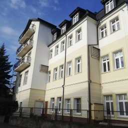 Eclectic Sp z o.o. - Odnowiona elewacja wielopiętrowego budynku z balkonami, pomalowana na kremowo i biało, z widoczną drabiną opartą o balkony i fragmentem rusztowania, na tle błękitnego nieba z chmurami i wysokim...