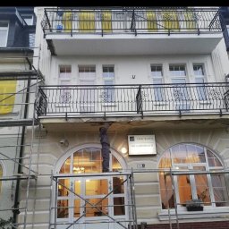 Eclectic Sp z o.o. - Pracownik na rusztowaniu naprawia uszkodzony balkon w kamienicy z ozdobnymi balkonami i oknami łukowymi, widoczny szyld firmy AMW Rewita DW Grunwald.