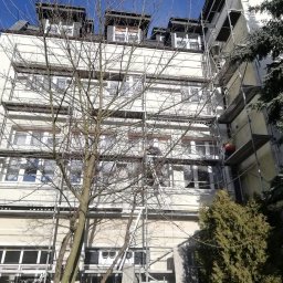 Eclectic Sp z o.o. - Wielopiętrowy budynek w trakcie prac elewacyjnych, z rusztowaniem i pracownikiem w pomarańczowej kamizelce na wysokości, widoczny przez gałęzie drzewa.