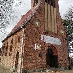 Eclectic Sp z o.o. - Ceglany kościół z czerwonym dachem, z banerem z wizerunkiem Jana Pawła II nad wejściem, trzema flagami po lewej i dużym drewnianym krzyżem po prawej stronie.