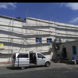 Eclectic Sp z o.o. - Budynek w trakcie ocieplania elewacji, widoczne rusztowania, częściowo otynkowana ściana, srebrny bus z otwartymi drzwiami i pracownik w niebieskiej koszulce przy wejściu do budynku.