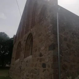 Fragment murowanej ściany budynku z kamienia i cegły z widocznymi oknami łukowymi i rynną spustową, w pochmurny dzień.