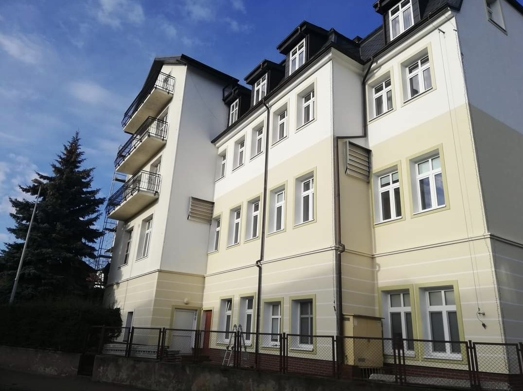 Odnowiona elewacja wielopiętrowego budynku z balkonami, pomalowana na kremowo i biało, z widoczną drabiną opartą o balkony i fragmentem rusztowania, na tle błękitnego nieba z chmurami i wysokim...