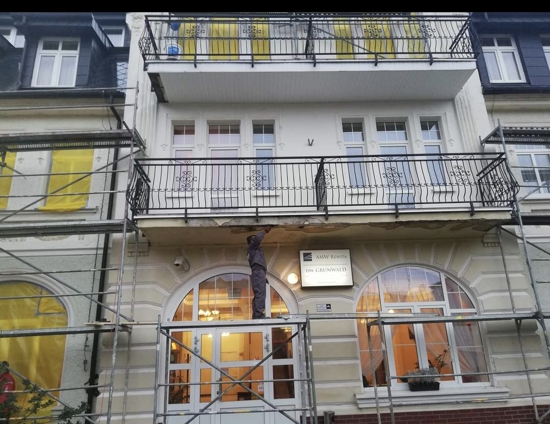 Pracownik na rusztowaniu naprawia uszkodzony balkon w kamienicy z ozdobnymi balkonami i oknami łukowymi, widoczny szyld firmy AMW Rewita DW Grunwald.