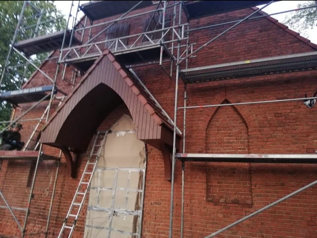 Renowacja ceglanej fasady budynku z charakterystycznymi gotyckimi łukami okiennymi, widoczne rusztowania i pracownik na rusztowaniu, okno zabezpieczone folią, fragment dachu z drewnianym podbitką.