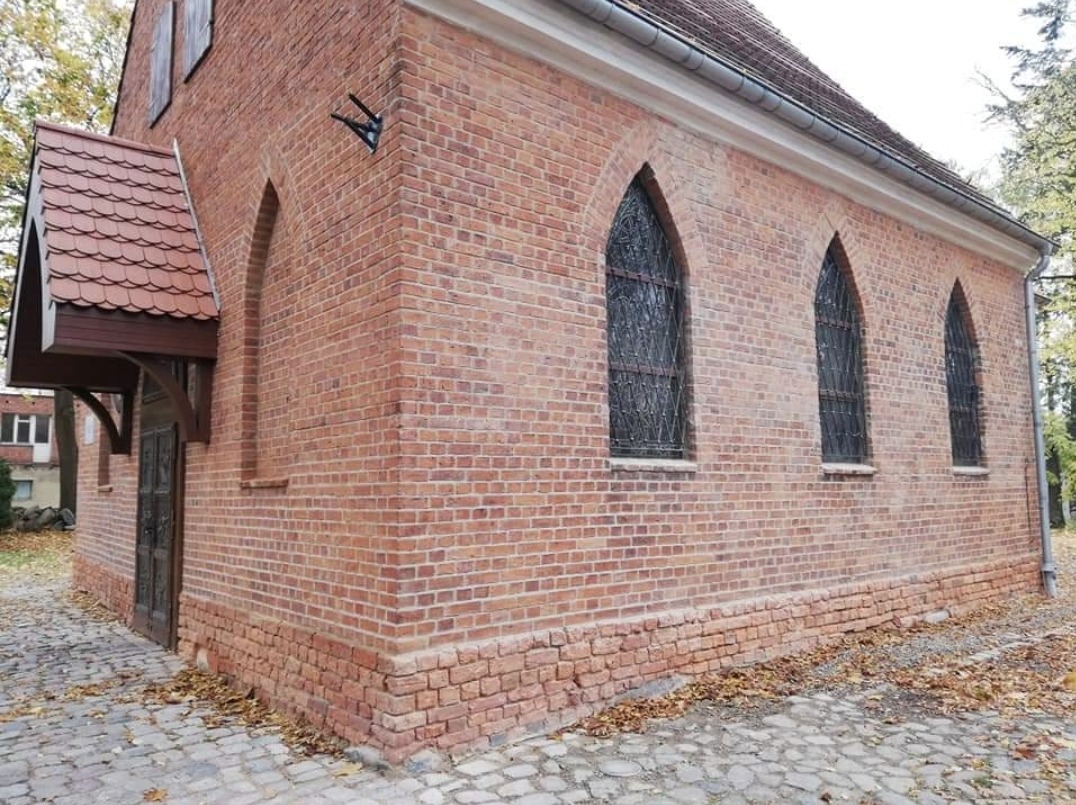 Zewnętrzny mur z czerwonej cegły z trzema ostrołukowymi oknami z dekoracyjnymi kratami, widoczny fragment dachu z dachówką i kamienny chodnik z opadłymi liśćmi.