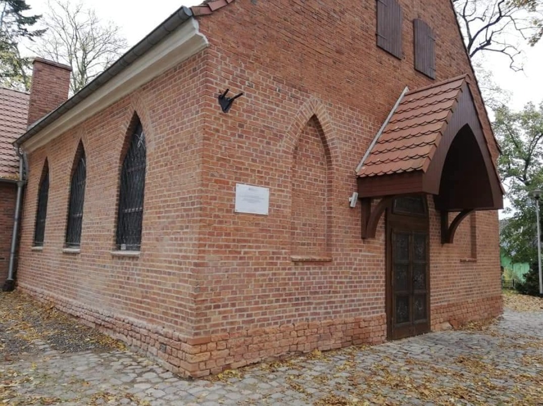 Ceglany budynek z gotyckimi oknami i drewnianym zadaszeniem nad wejściem, widoczne detale architektoniczne i brukowa nawierzchnia.