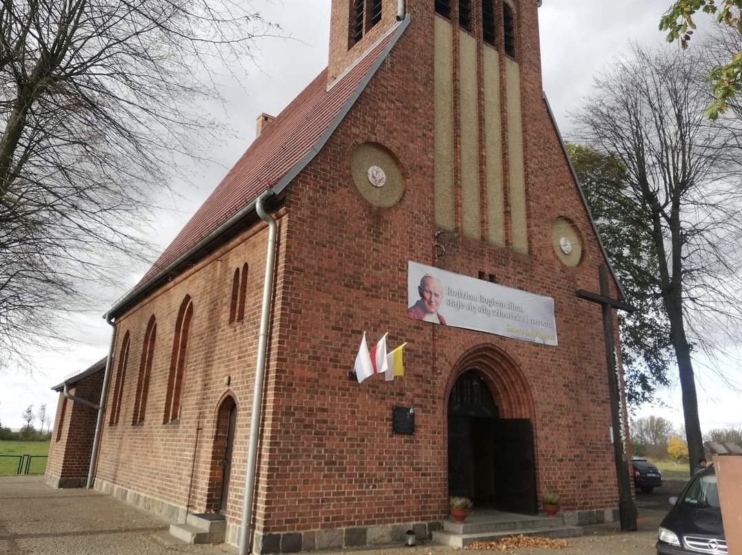 Ceglany kościół z czerwonym dachem, z banerem z wizerunkiem Jana Pawła II nad wejściem, trzema flagami po lewej i dużym drewnianym krzyżem po prawej stronie.