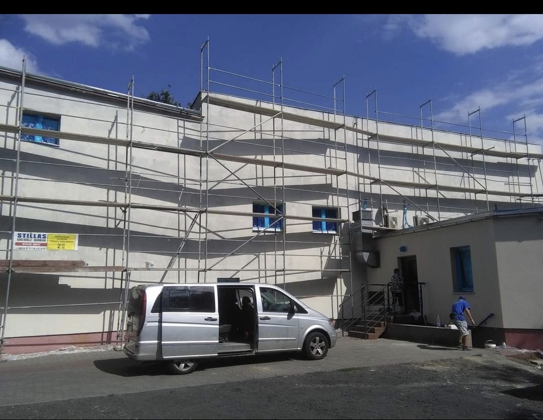 Budynek w trakcie ocieplania elewacji, widoczne rusztowania, częściowo otynkowana ściana, srebrny bus z otwartymi drzwiami i pracownik w niebieskiej koszulce przy wejściu do budynku.