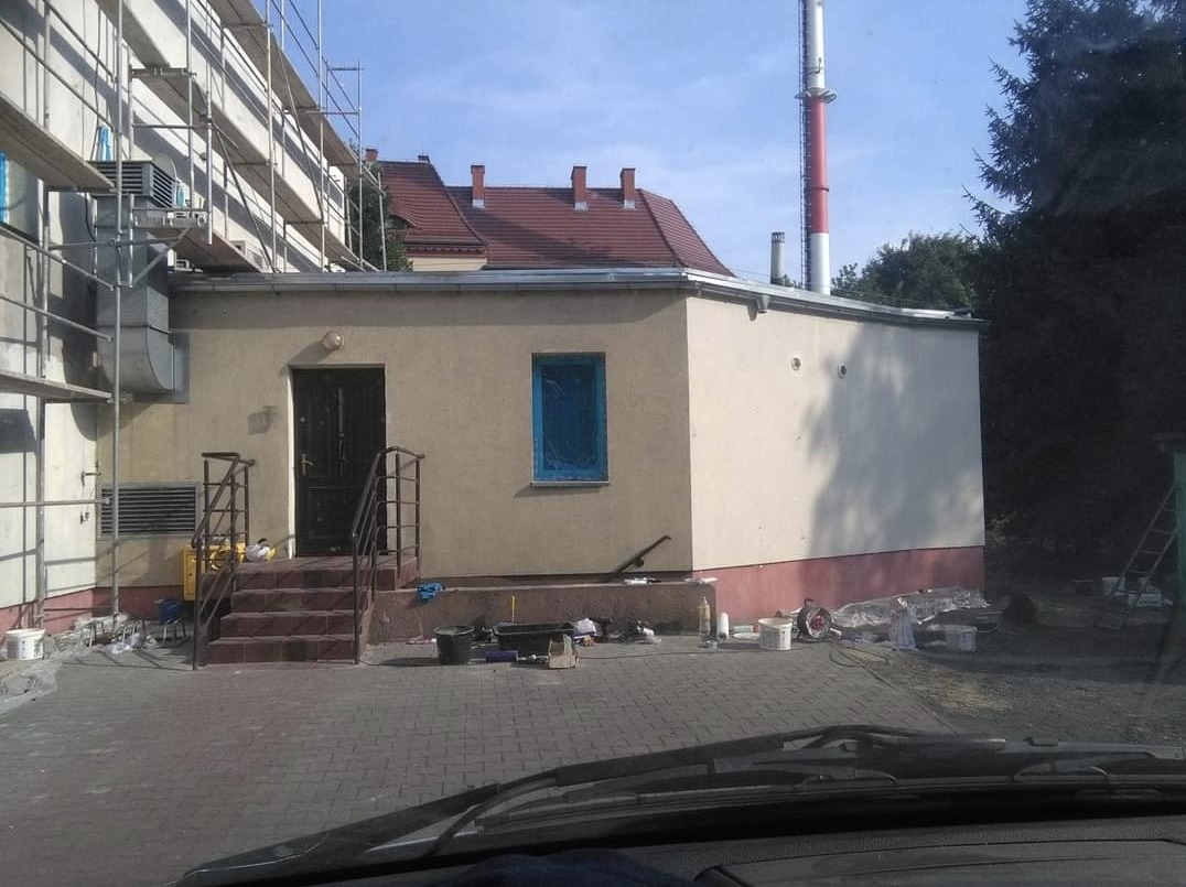 Budynek w trakcie ocieplania, z widocznymi rusztowaniami, oknem zabezpieczonym folią, schodami wejściowymi i rozłożonymi narzędziami malarskimi na kostce brukowej.