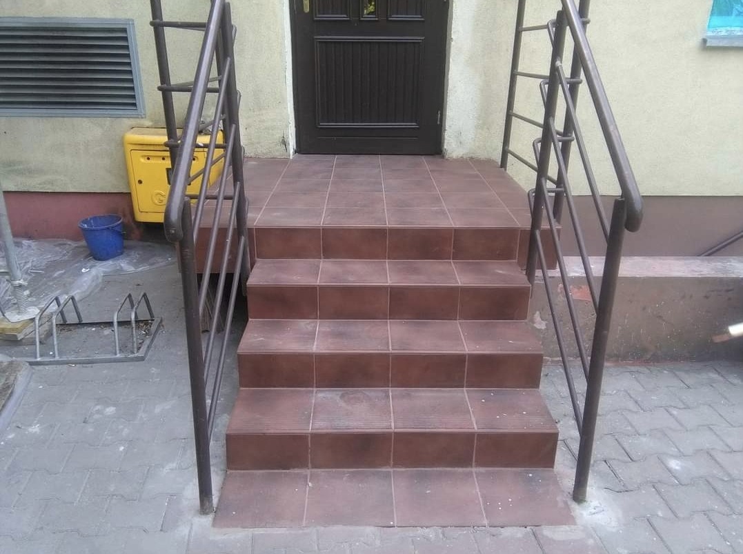 Wykończone schody wejściowe do budynku, wyłożone brązowymi płytkami ceramicznymi, z metalową barierką i żółtą skrzynką gazową w tle.