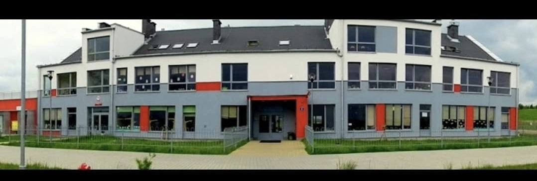 Dwukondygnacyjny budynek szkoły pomalowany na biało, szaro i z czerwonymi akcentami, z ciemnym dachem i licznymi oknami, otoczony niskim płotkiem i zielenią.