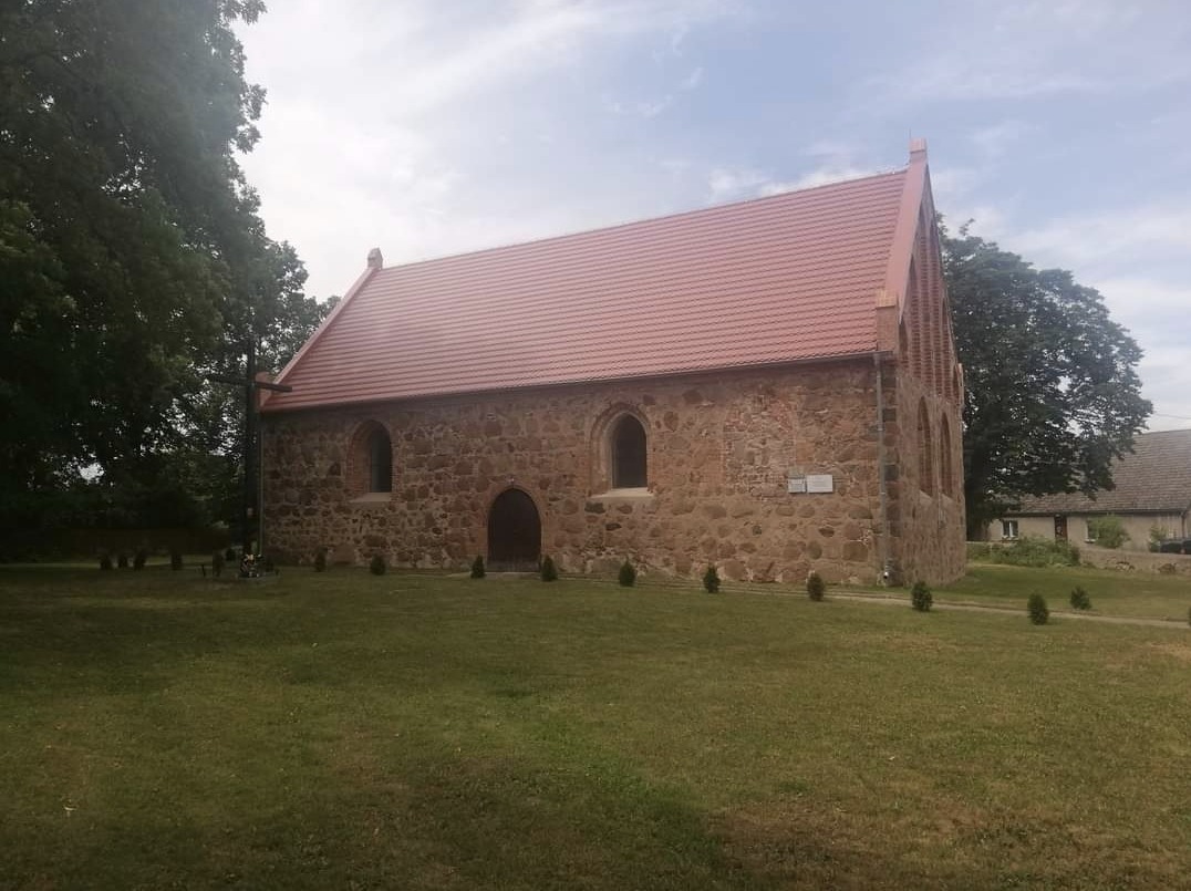 Kamienny budynek kościoła z czerwoną dachówką, widok z zewnątrz na tle zielonego trawnika i drzew.