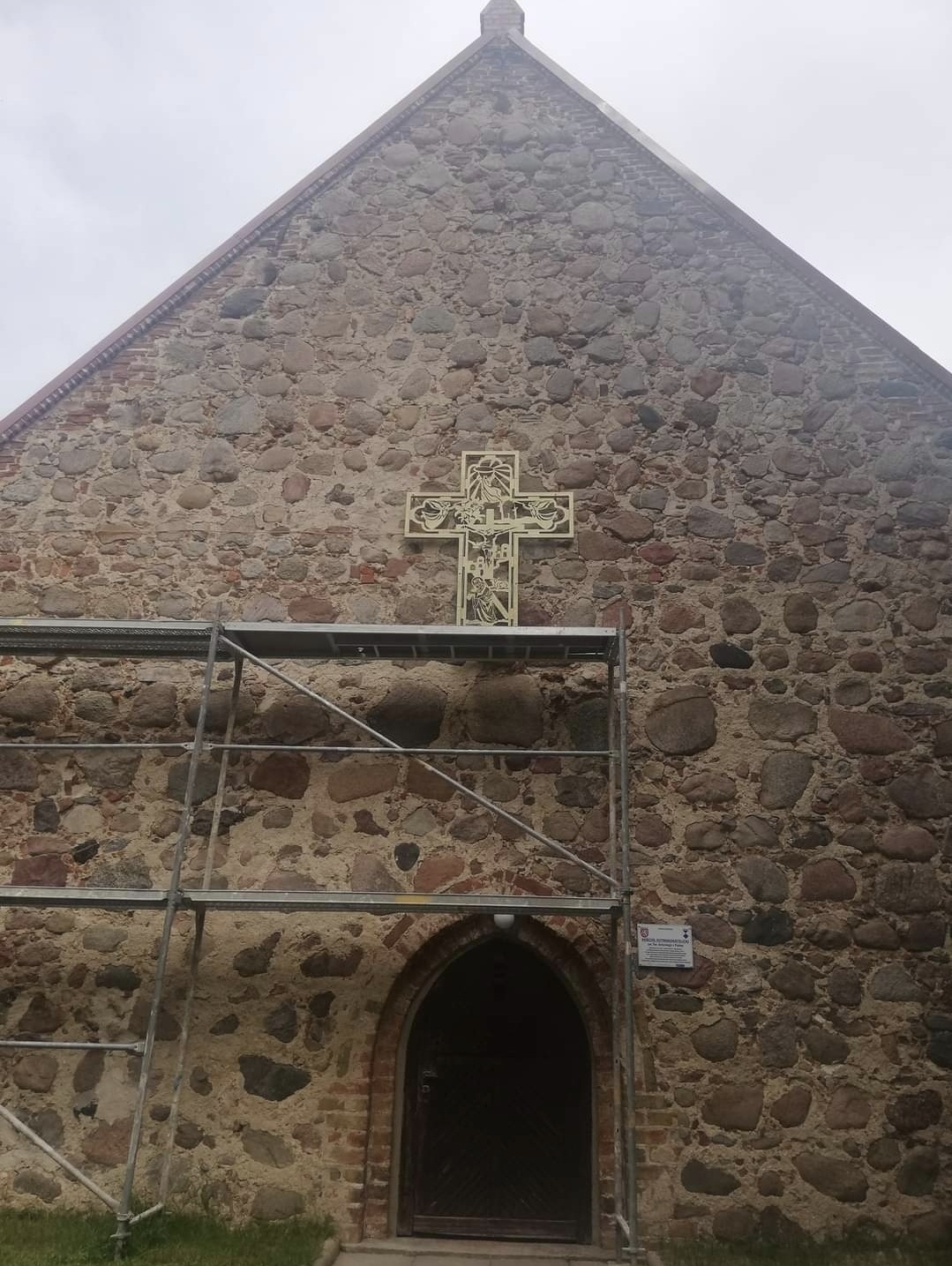 Kamienna fasada kościoła z ozdobnym krzyżem, częściowo zasłonięta rusztowaniem, widoczny fragment łukowego wejścia.