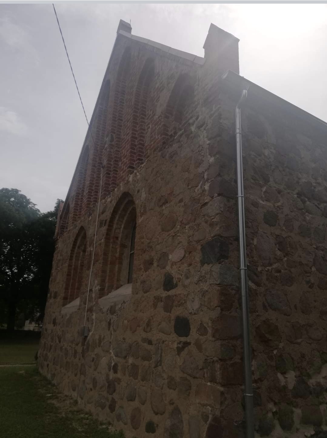 Fragment murowanej ściany budynku z kamienia i cegły z widocznymi oknami łukowymi i rynną spustową, w pochmurny dzień.