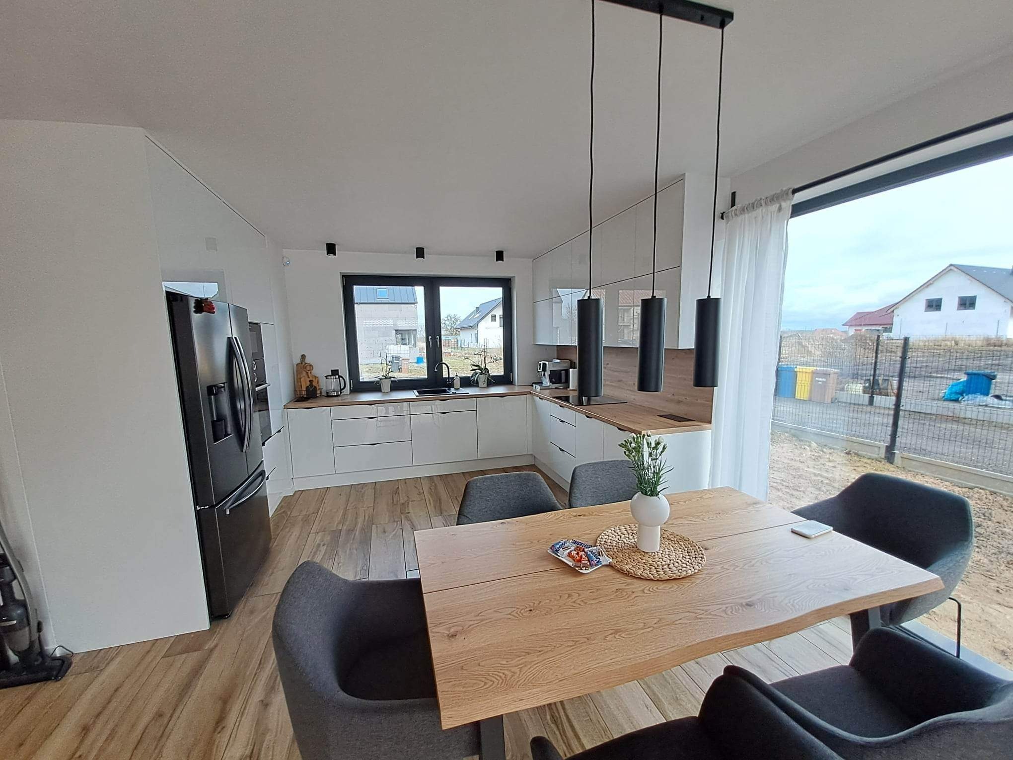 Nowoczesna, jasna kuchnia z drewnianym blatem i stołem w Szczecinie. Minimalistyczne białe szafki, czarne lampy wiszące i podłoga imitująca drewno tworzą spójną całość. Na stole dekoracja...