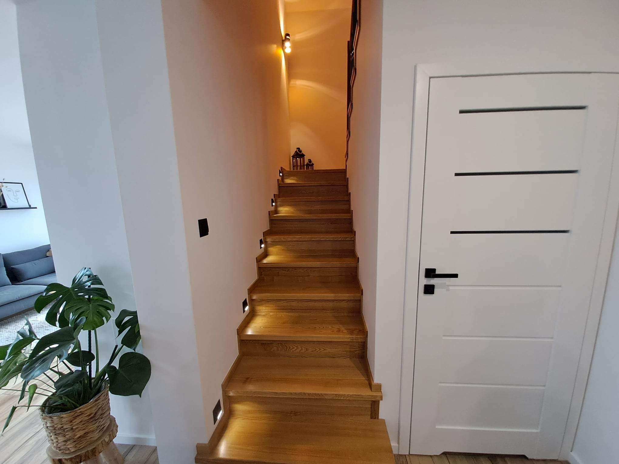 Drewniane schody z podświetleniem LED wzdłuż ściany, prowadzące na wyższą kondygnację, obok białe drzwi z poziomymi wgłębieniami i czarną klamką, oraz roślina Monstera w plecionym koszu.
