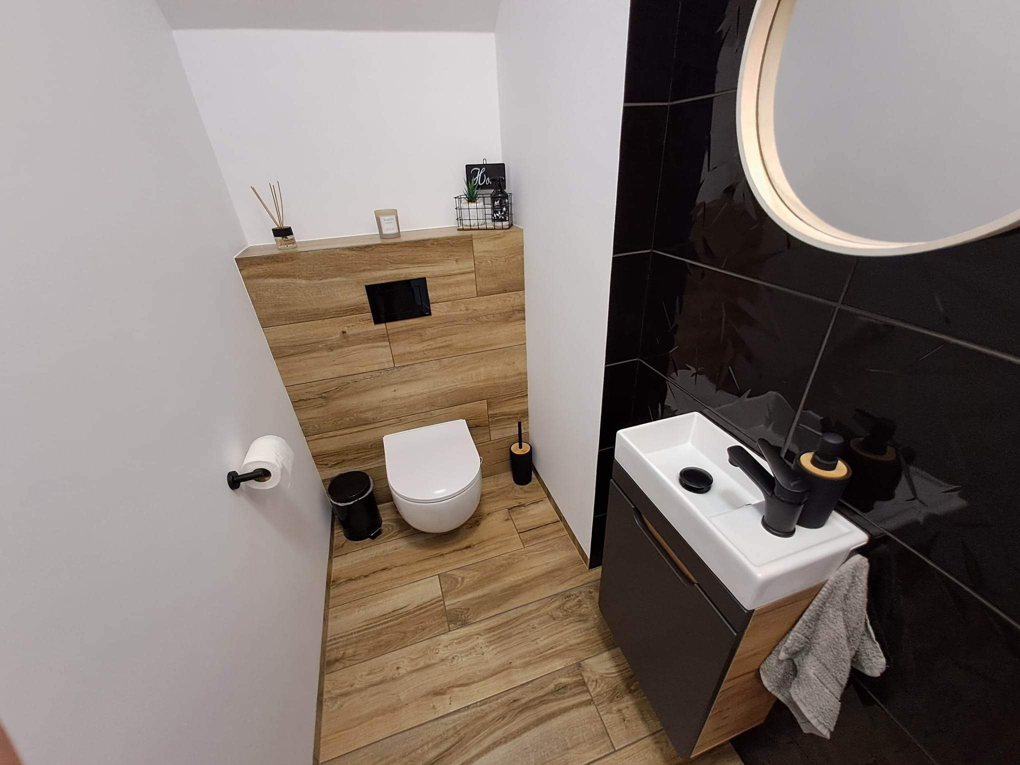 Nowoczesna toaleta z drewnopodobną podłogą i okładziną, czarne płytki strukturalne na ścianie, biała ceramika sanitarna, minimalistyczna umywalka z czarną armaturą i okrągłe podświetlane lustro.