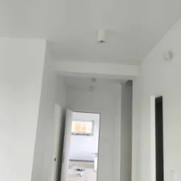 Świeżo pomalowany na biało korytarz z widocznym oświetleniem sufitowym, otwartymi drzwiami i zabezpieczoną podłogą w pomieszczeniu obok.