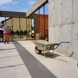 Betonowa konstrukcja w trakcie budowy z deskowaniem, widoczna taczka z zaprawą, pracownik w pomarańczowych spodenkach i betonowa ściana z otworami po szalunkach.