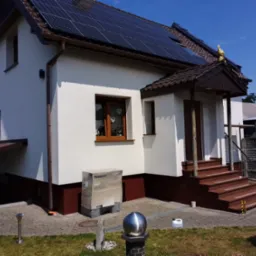 Biały dom jednorodzinny z brązowymi elementami wykończeniowymi, panelami słonecznymi na dachu, ozdobną figurką koguta na szczycie dachu, schodami wejściowymi wyłożonymi brązowym kamieniem,...