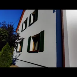 Elewacja budynku z białym tynkiem, zielonymi okiennicami i brązową rynną, widoczna z perspektywy żabiej, na tle błękitnego nieba i zieleni drzew.