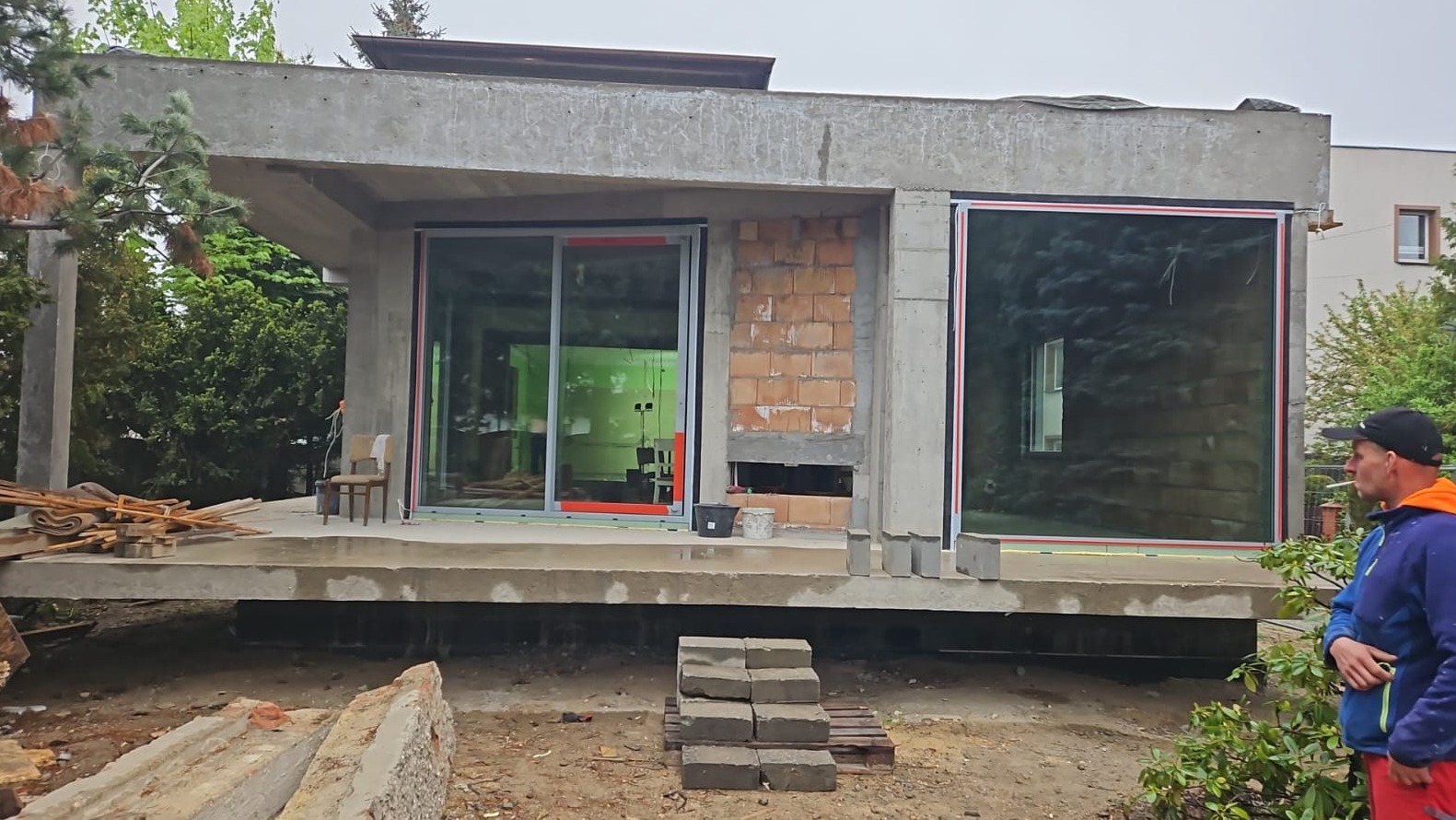 Surowy stan budynku żelbetowego z dużymi oknami i fragmentem murowanej ściany. W tle zieleń, na pierwszym planie stos cegieł i mężczyzna w roboczym ubraniu.