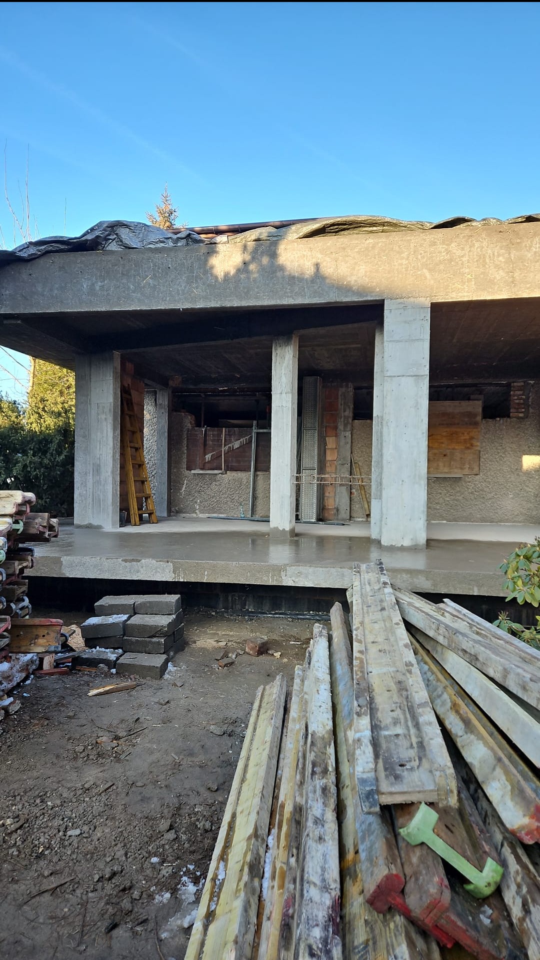 Surowa konstrukcja budynku w trakcie budowy z widocznymi betonowymi słupami, deskami szalunkowymi i elementami konstrukcyjnymi. Widok na plac budowy w słoneczny dzień.