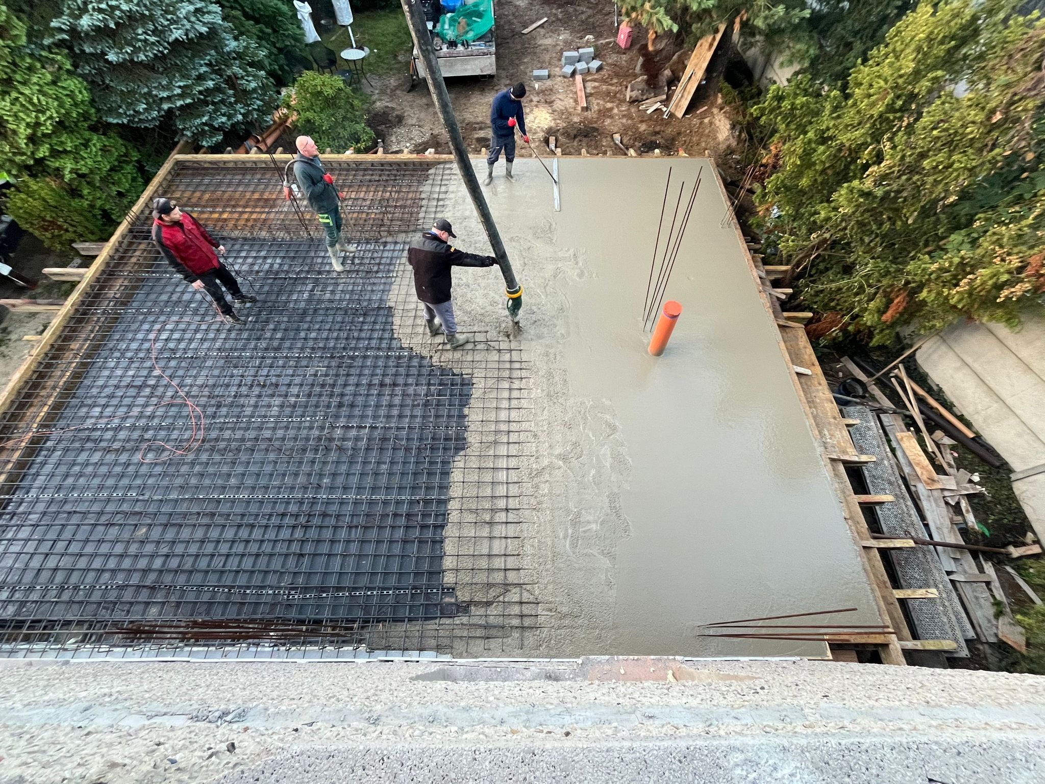 Wylewka betonu na zbrojenie stropu z użyciem pompy, widoczne pręty zbrojeniowe i pracownicy. Częściowo zalana powierzchnia. Ujęcie z góry.