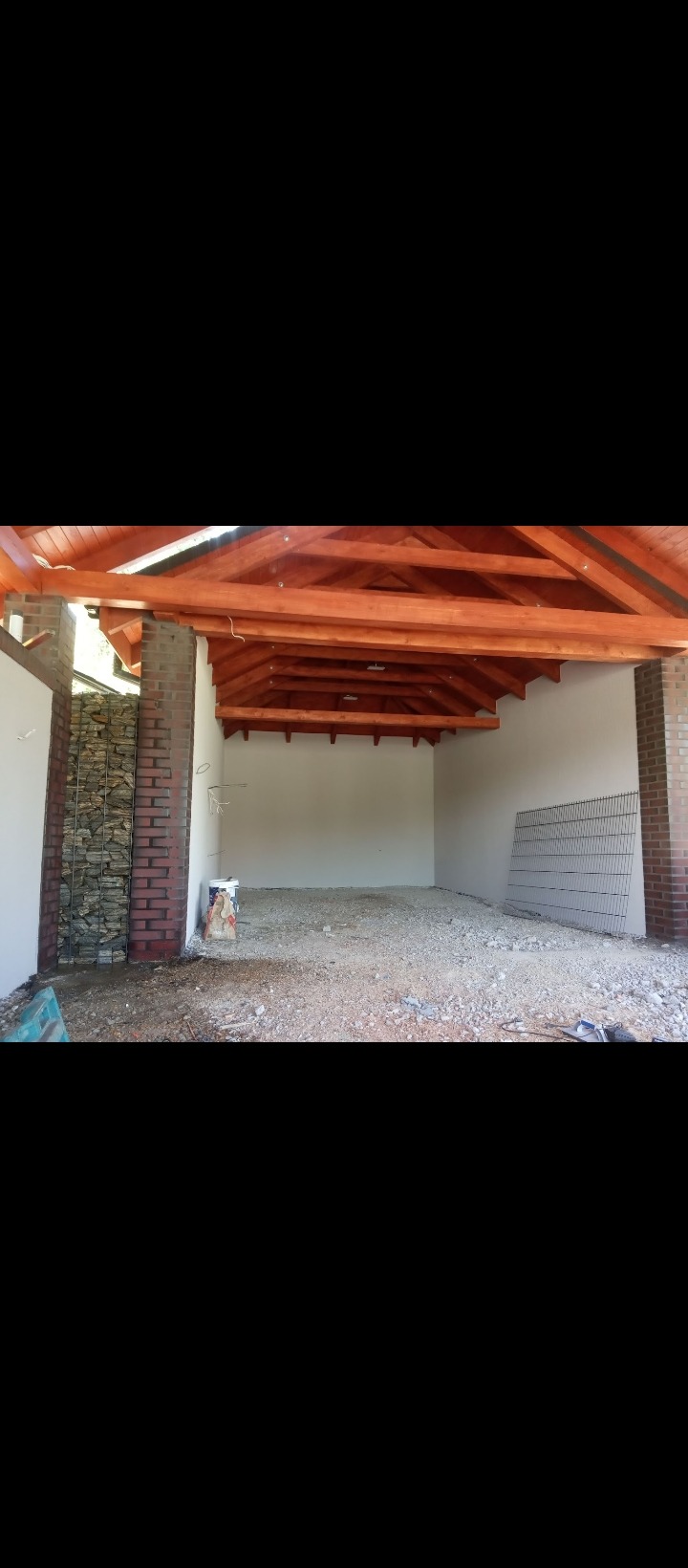 Wnętrze wiaty z drewnianą konstrukcją dachu, ceglanymi filarami z elementami gabionowymi, przygotowane pod wylewkę, widoczny piasek i siatka zbrojeniowa.