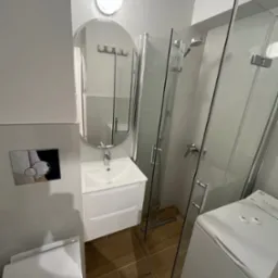 Nowoczesna łazienka w odcieniach szarości i bieli z brązowymi płytkami podłogowymi, owalnym lustrem nad białą szafką z umywalką, kabiną prysznicową ze szklanymi drzwiami oraz pralką.