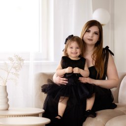 Ewelina Kwiatek- Fotografia - Portret matki z córką w pasujących czarnych sukienkach, siedzących na beżowej sofie w jasnym wnętrzu, z dekoracyjnym bukietem w wazonie na okrągłym stoliku.