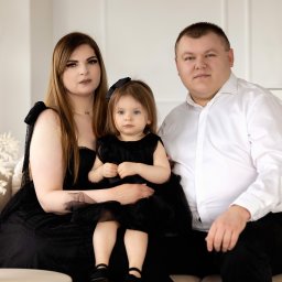Ewelina Kwiatek- Fotografia - Portret rodzinny: rodzice i córka w czarnych strojach siedzą na beżowej kanapie w jasnym wnętrzu z dekoracjami w tle.