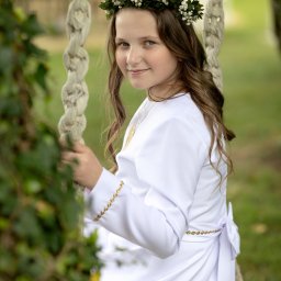 Ewelina Kwiatek- Fotografia - Dziewczynka w białej albie i wianku na głowie siedzi na huśtawce linowej w ogrodzie, spoglądając w obiektyw z delikatnym uśmiechem. Ujęcie portretowe, plener.