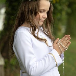 Ewelina Kwiatek- Fotografia - Dziewczynka w białej albie i wianku z kwiatów, z różańcem w dłoniach, spogląda w dół. Delikatne światło podkreśla detale stroju i skupienie na twarzy.