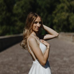 Ewelina Kwiatek- Fotografia - Portret kobiety w białej sukience na tle zieleni i kamiennego mostu. Ujęcie z boku, podkreślające delikatność i elegancję. Jasne, naturalne światło.