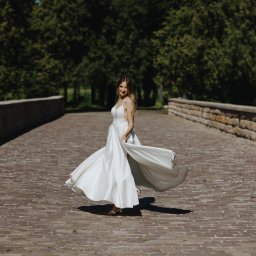 Ewelina Kwiatek- Fotografia - Uśmiechnięta panna młoda w białej sukni wiruje na kamiennym moście, otoczona zielenią drzew. Słoneczny dzień podkreśla blask sukni i radość chwili.