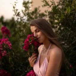 Kobieta w satynowej sukience wącha bukiet różowych kwiatów na tle zieleni i beżowego budynku w ciepłym świetle zachodzącego słońca.