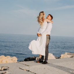 Ewelina Kwiatek- Fotografia - Para młoda na betonowym nabrzeżu z widokiem na morze; mężczyzna trzyma kobietę w białej sukni w ramionach, oboje patrzą na siebie z uśmiechem.