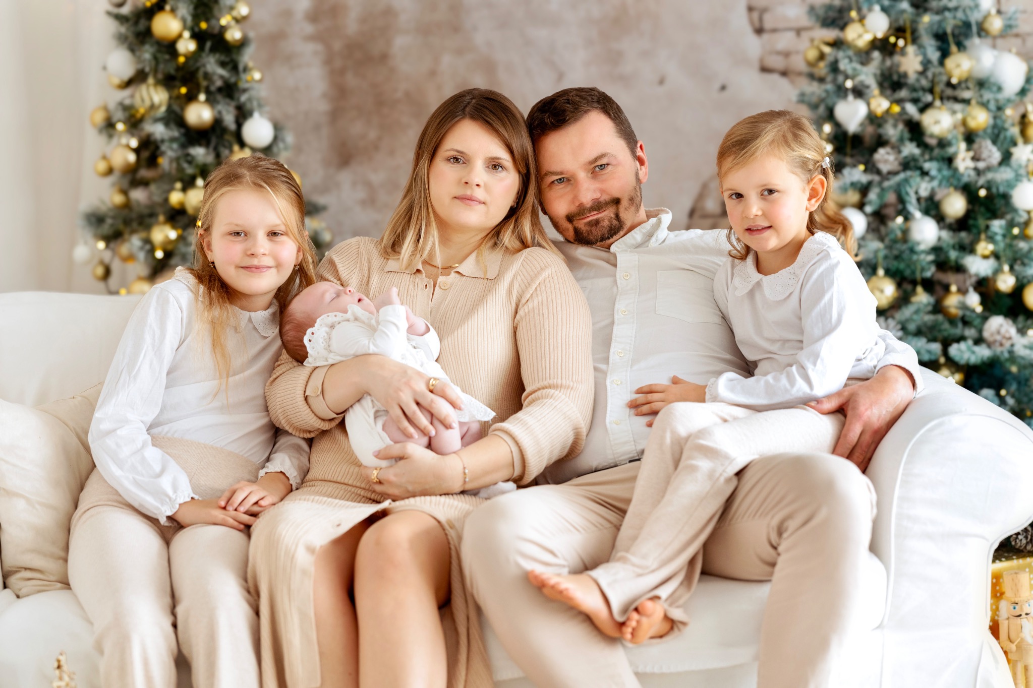 Rodzinne zdjęcie w świątecznej scenerii: rodzice z dwiema córkami i niemowlęciem, wszyscy ubrani w jasne, kremowe kolory, siedzą na białej sofie przed choinkami ozdobionymi złotymi i białymi bombkami.