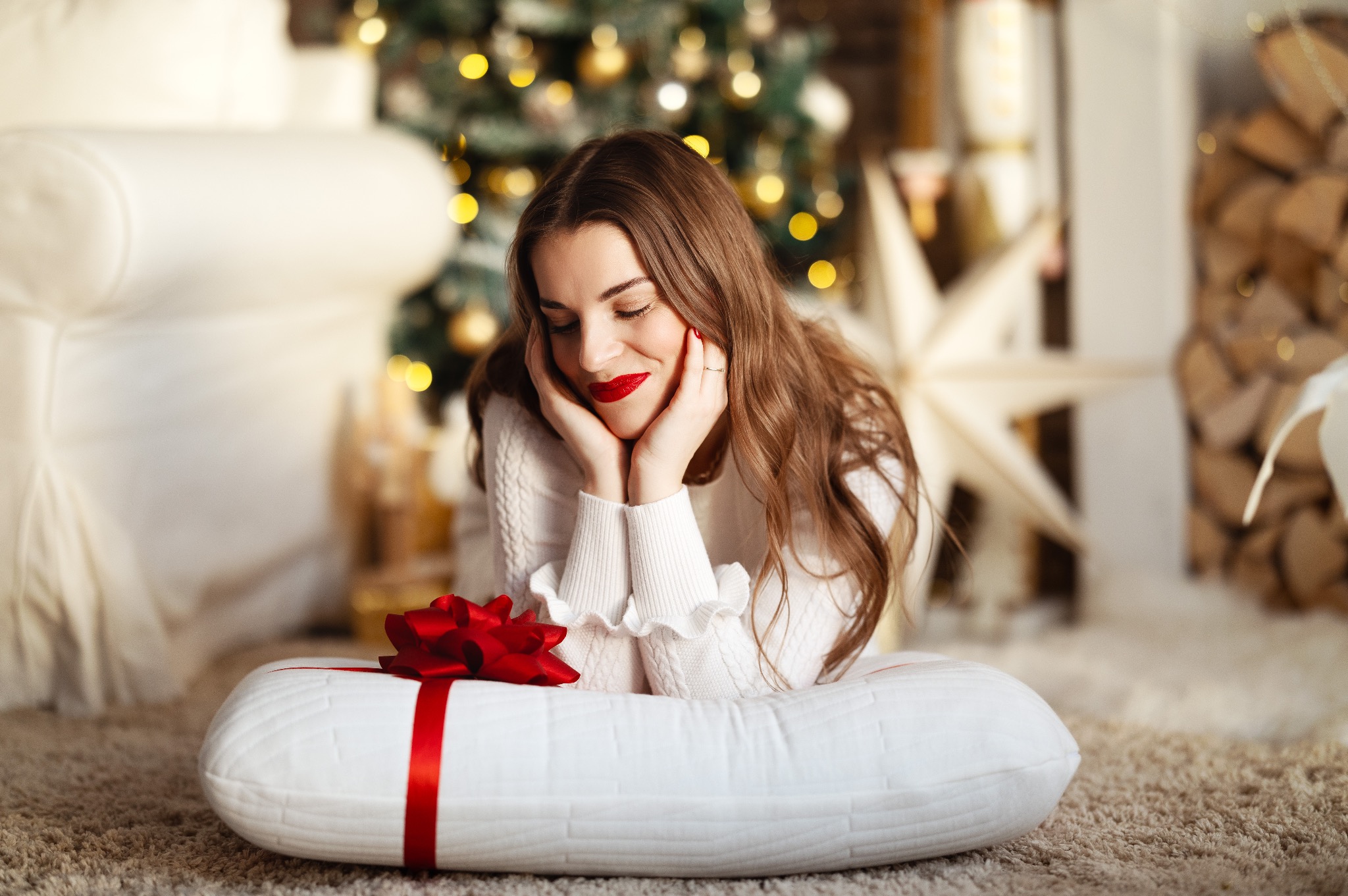 Uśmiechnięta brunetka z czerwonymi ustami leży na poduszce z czerwoną kokardą, w tle choinka z lampkami i drewno kominkowe.