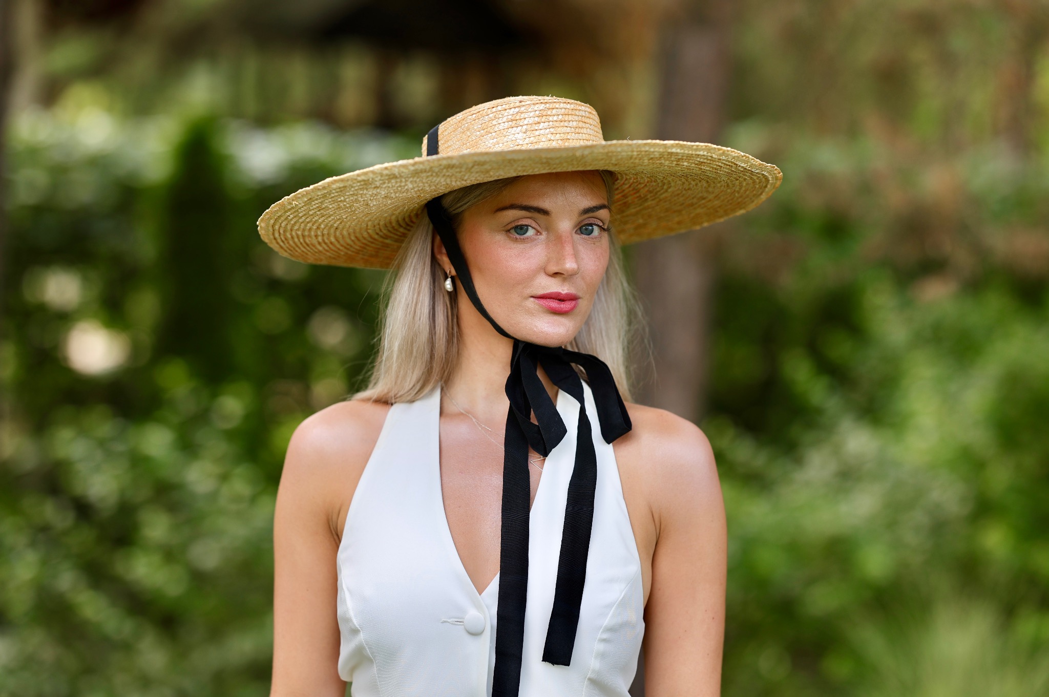Portret kobiety w słomkowym kapeluszu z czarną wstążką, białej sukience na tle zieleni.