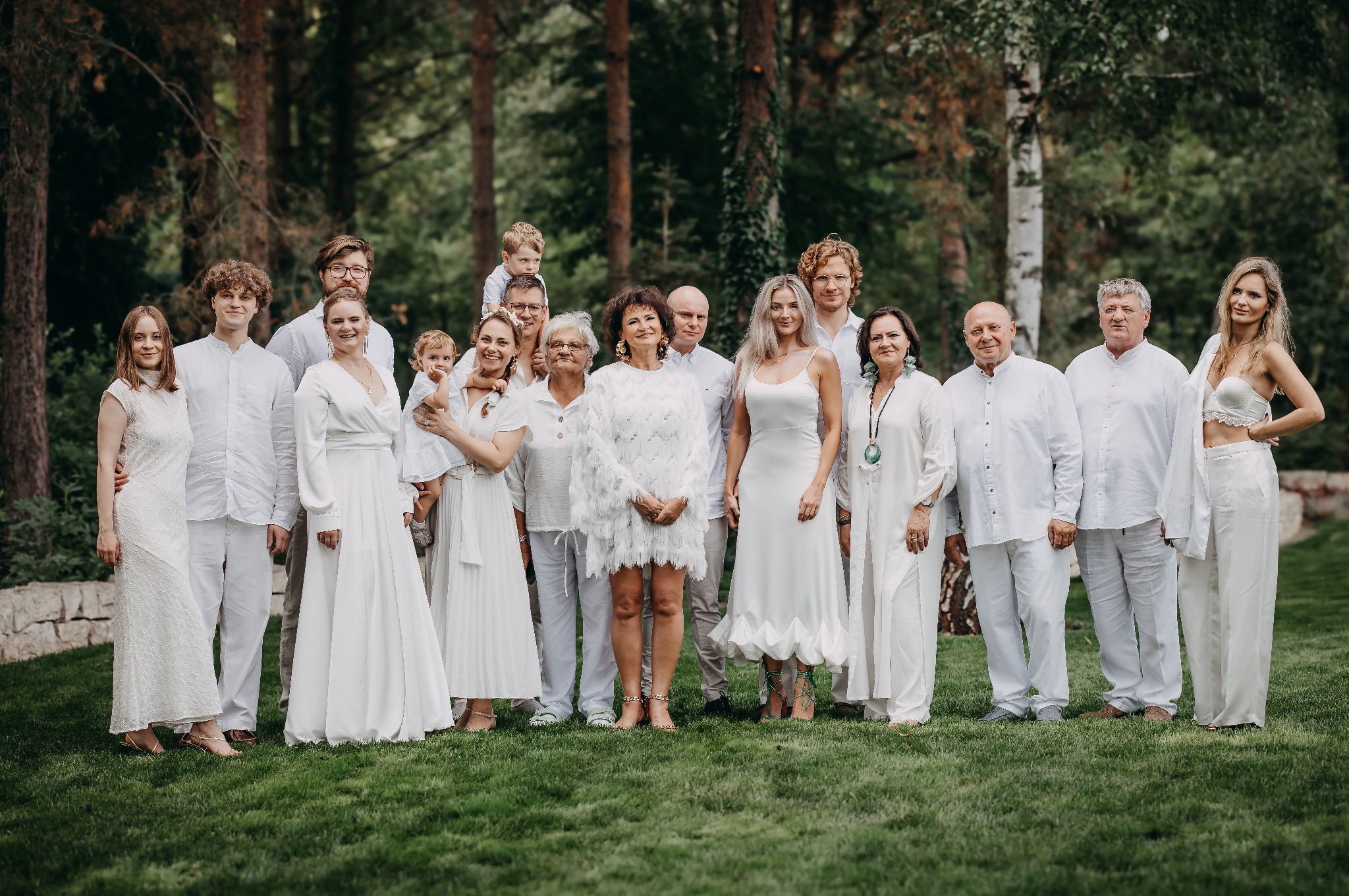 Duża grupa ludzi ubranych na biało pozuje na trawniku przed lasem, różnorodne style ubioru i wieku, uśmiechnięte twarze, swobodna atmosfera.