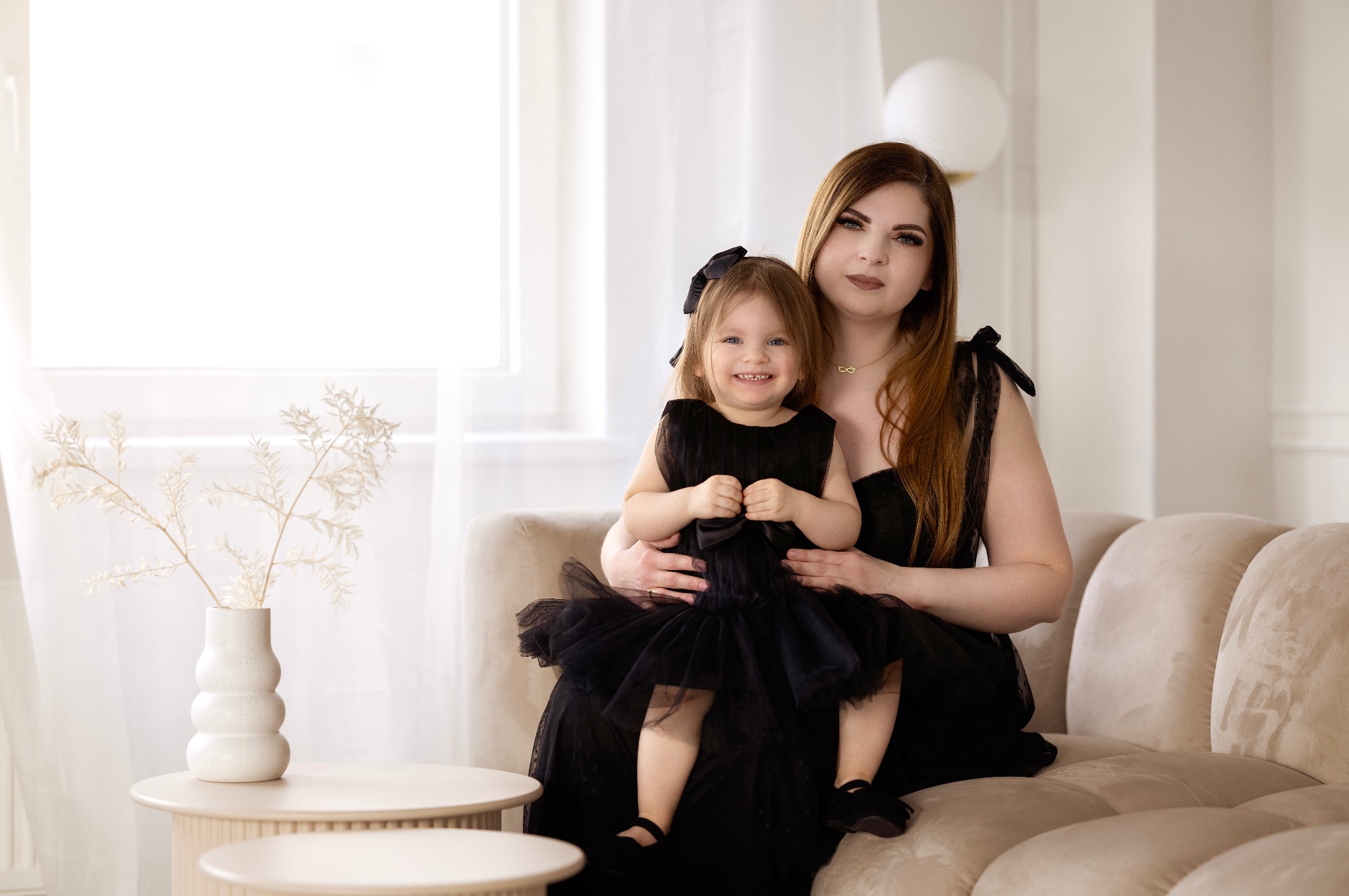 Portret matki z córką w pasujących czarnych sukienkach, siedzących na beżowej sofie w jasnym wnętrzu, z dekoracyjnym bukietem w wazonie na okrągłym stoliku.