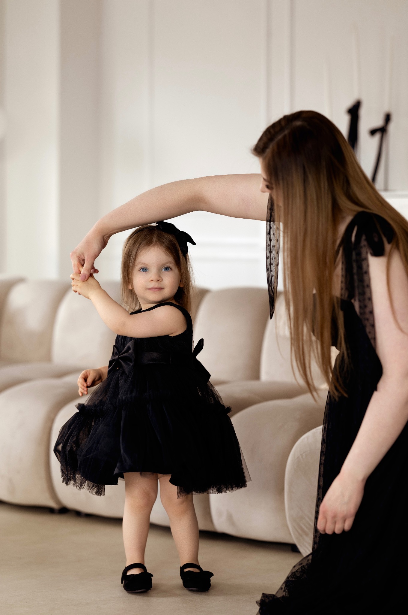 Matka trzymająca za rękę córeczkę w czarnej, tiulowej sukience z kokardą, na tle jasnej sofy w minimalistycznym wnętrzu.