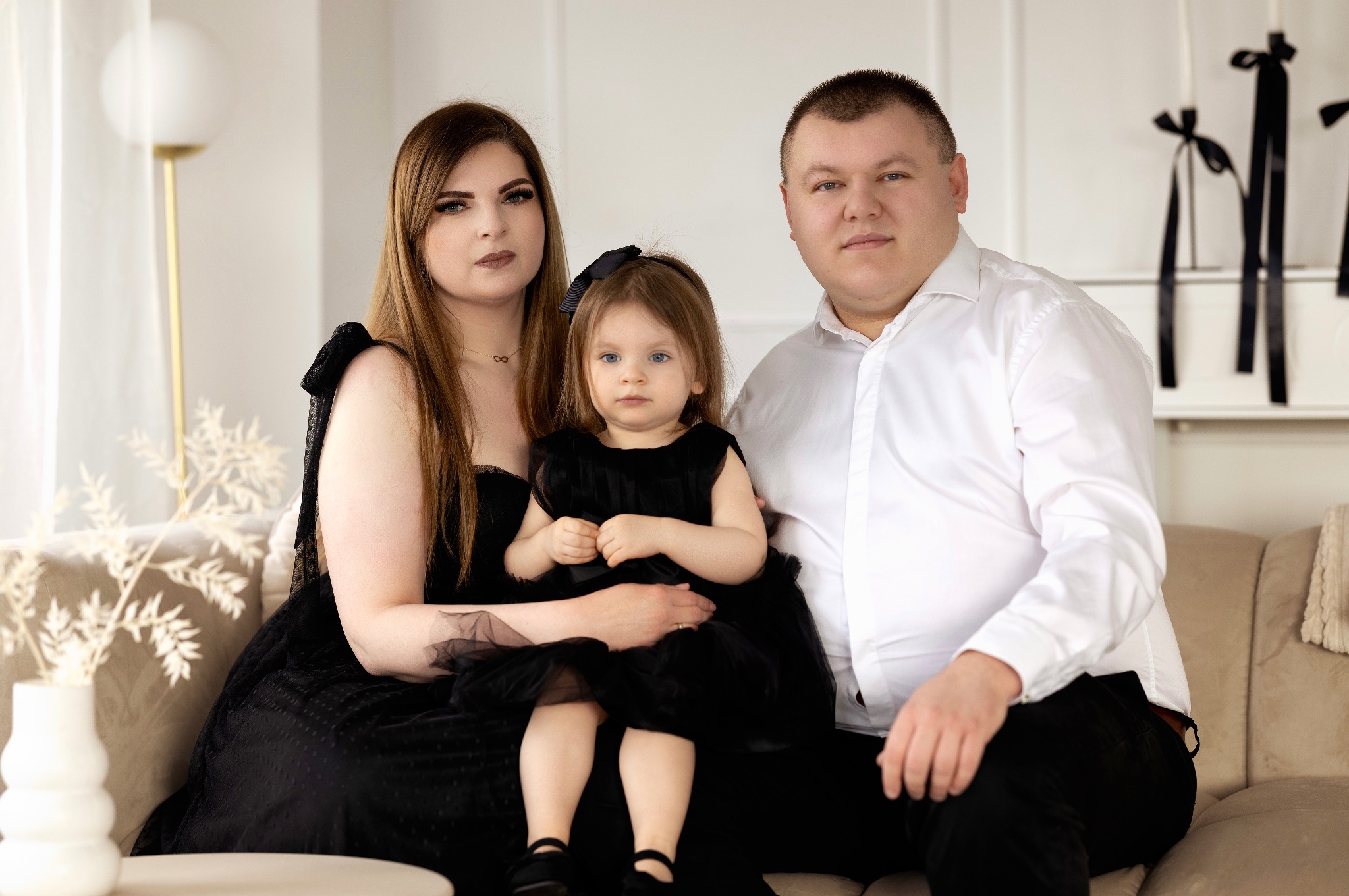 Portret rodzinny: rodzice i córka w czarnych strojach siedzą na beżowej kanapie w jasnym wnętrzu z dekoracjami w tle.