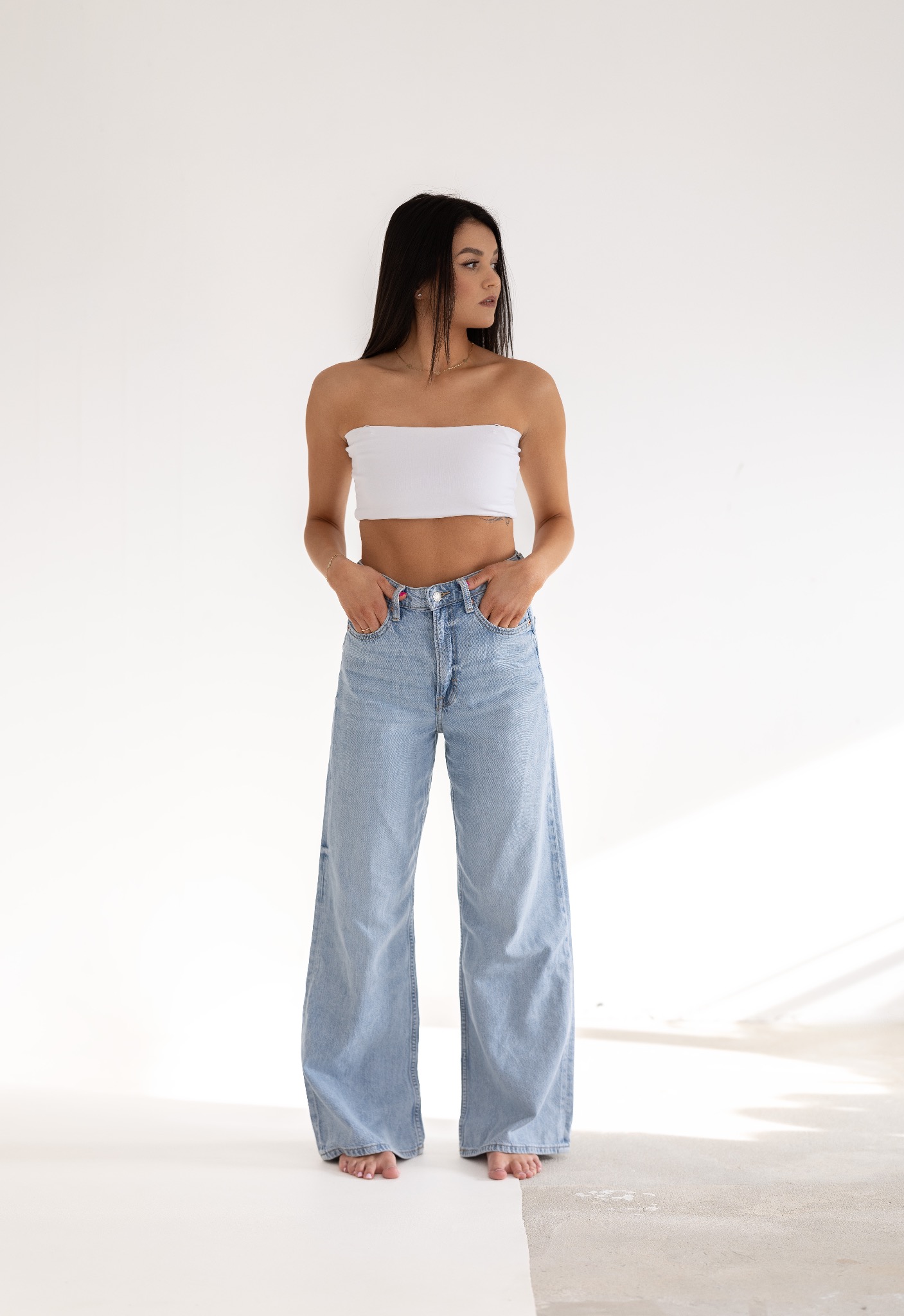 Portret kobiety w białym topie i szerokich jeansach na jasnym tle, z rękami w kieszeniach i spojrzeniem skierowanym w bok.