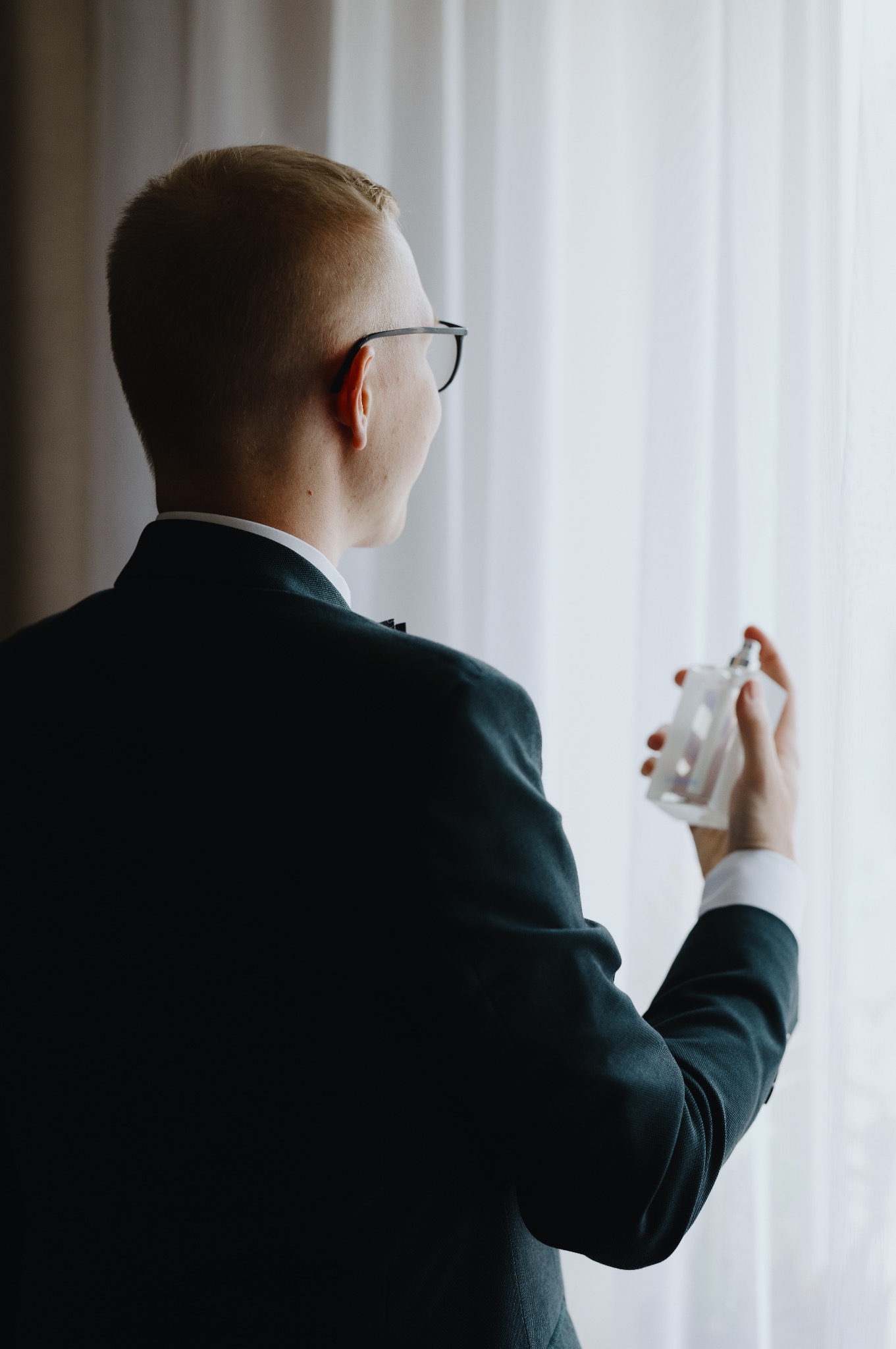 Mężczyzna w garniturze, z tyłu, spryskuje się perfumami przed oknem z delikatnymi zasłonami. Przygotowania do uroczystości, elegancki strój.